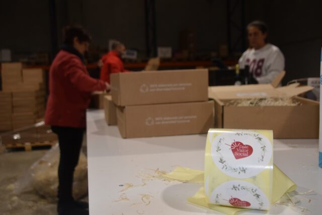 Imagen. Primer plano del packaging personalizado de los lotes de Navidad para empresas de más de 50 trabajadores del proyecto 'Dale Valor Social' que ha impulsado la Fundación AltoAragón Integración (FAI) a través de Somontano Social, su Centro Especial de Empleo, y al fondo trabajadores preparando los pedidos.