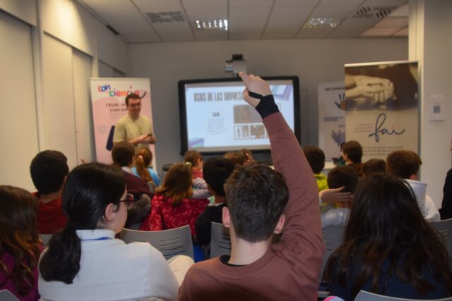 Imagen. Foto de una clase, con los alumnos de espaldas. En primer término, un alumno levanta la mano para preguntar al docente dentro del proyecto Con-Ciencia.