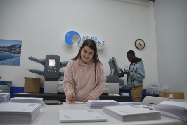 Imagen. En el centro de la imagen, Lucía, alumna del programa experiencial Valentia Documenta II, durante sus prácticas en el departamento de Preimpresión y Ensobrado de Somontano Social, el Centro Especial de Empleo de la Fundación AltoAragón Integración (FAI). Está en una mesa, junto a varios montones de sobres. Al fondo, la máquina de ensobrar.