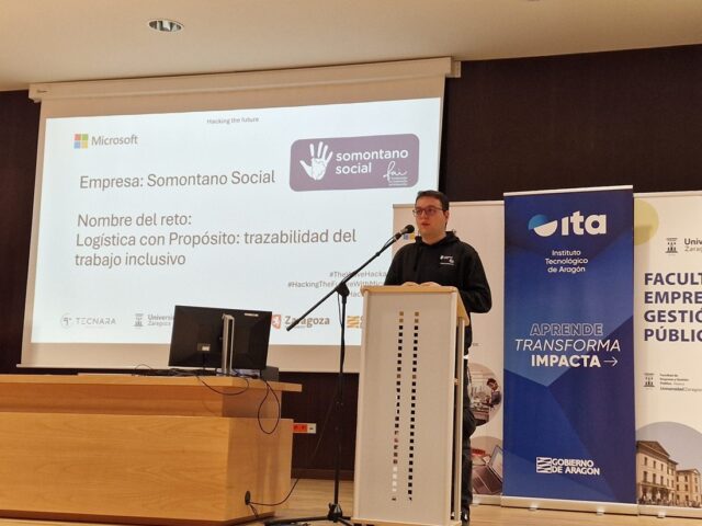 Imagen. Ángel Salamero, detrás de un atril a la derecha de la imagen, detalla el reto propuesto por Somontano Social en el Hackathon The Wave Huesca 2026.