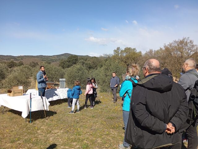 Imagen. Al fondo de la imagen, un guía está realizando una explicación sobre las abejas junto a unas colmenas vacías situadas sobre dos mesas. Alrededor, de espaldas, un grupo de trabajadores de Somontano Social y sus familias, participantes en la ruta interpretativa organizada por el Centro Especial de Empleo de la Fundación AltoAragón Integración (FAI).
