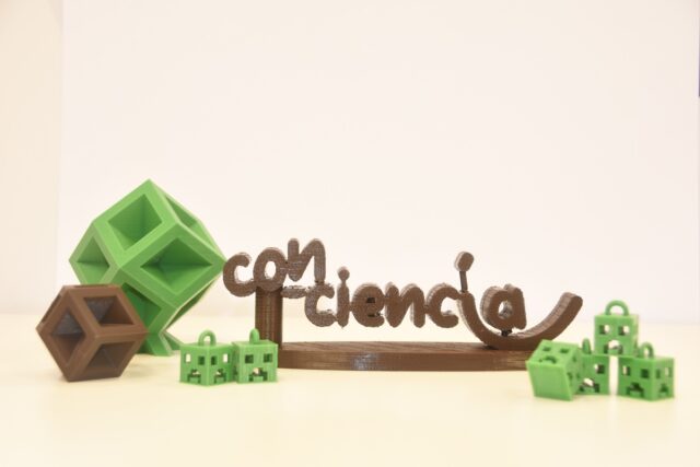 Imagen. Logotipo de Con-Ciencia realizado en impresión 3D con filamento de color marrón, rodeado por otros objetos realizados con fabricación aditiva en filamento de color verde, como varios creeper del juego Minecraft.