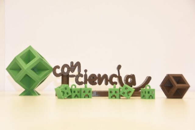 Imagen. Logotipo de Con-Ciencia realizado en impresión 3D con filamento de color marrón, rodeado por otros objetos realizados con fabricación aditiva en filamento de color verde, como varios creeper del juego Minecraft.
